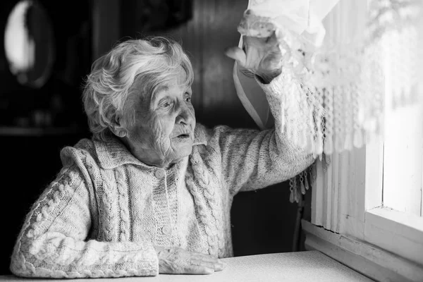 Siyah ve beyaz fotoğraf yaşlı bir kadın, evin içinde belgili tanımlık pencere perde kaldırdı.