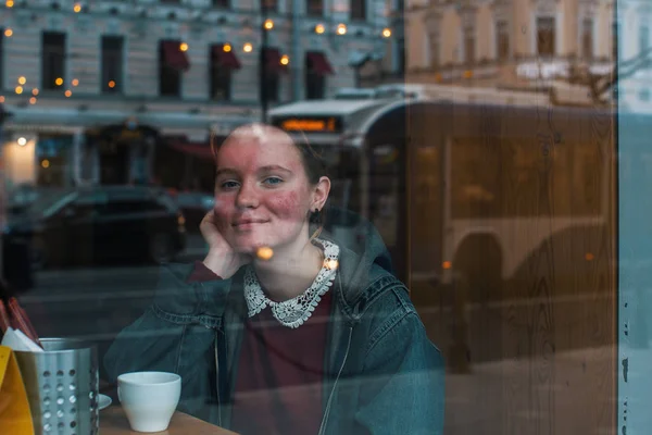 ウィンドウで市の光が反射して 通りを見て カフェに座ってコーヒーのカップを持つ少女 — ストック写真