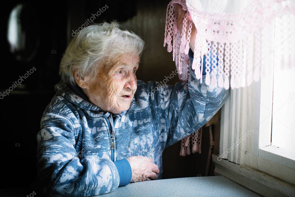 An elderly woman raising the curtain looks out the window.