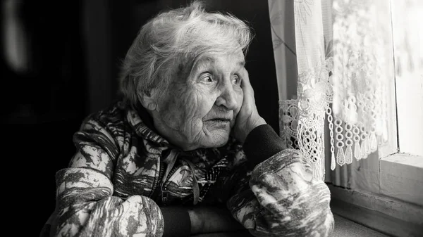 老妇人在家里坐在桌子前望着窗外 单色照片 免版税图库照片