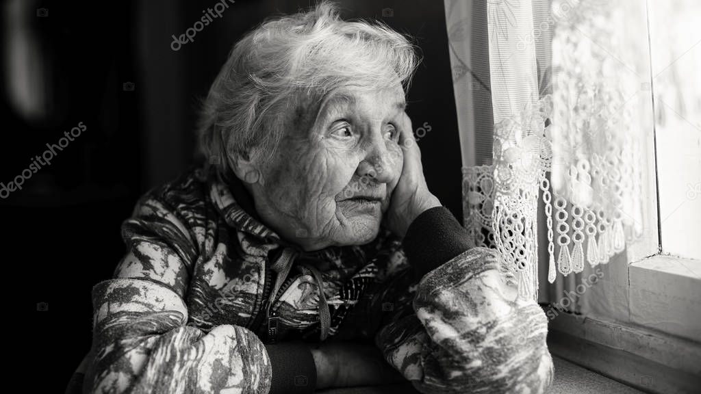 Elderly woman in the house sitting at the table looking out the window. Monochrome photo.