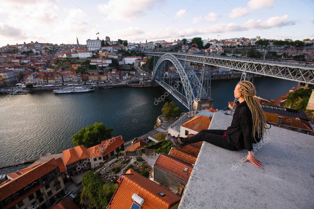 Young woman with blond dreadlocks sitting in front of Douro river and Dom luis I bridge in Porto, Portugal.