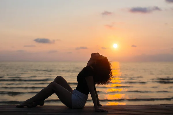 日落时坐在海边的年轻女子剪影 — 图库照片