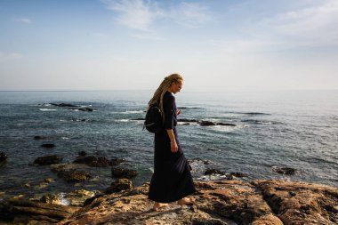 Okyanusa bakarak kayaların üzerinde duran sarışın dreadlocks ile genç bir kadın.