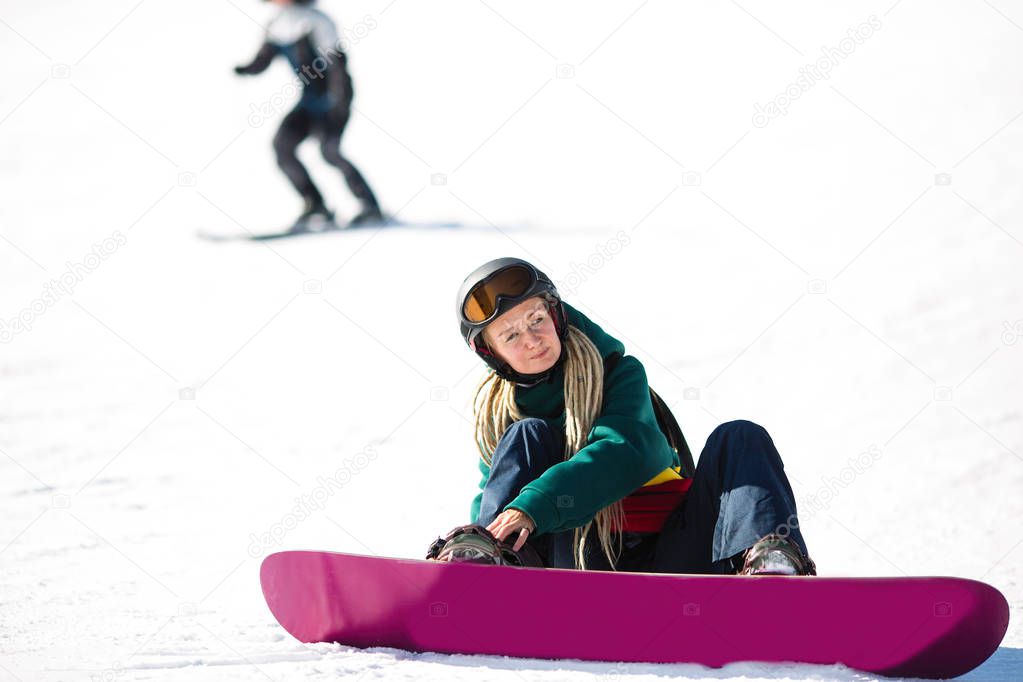 Young woman snowboarder with dreadlocks sitting zipping up his boots while sitting on a snowy slope.
