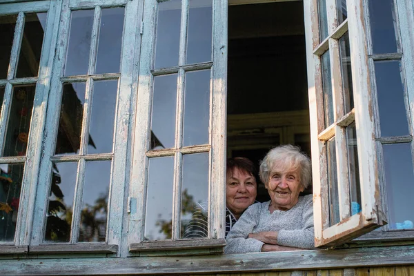 一位老妇人带着她成年的女儿看着村屋的窗户 — 图库照片