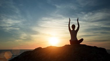 Yoga silueti gibi genç bir kadın okyanus sahilinde gün batımında egzersiz yapıyor..