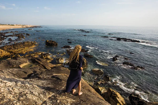 長い光のドレッドロックを持つ若い女性は サーフィンを見て海の岩の海岸に立っています — ストック写真