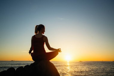 Fantastik gün batımında okyanus sahilinde yoga yapan siluet genç kadın.