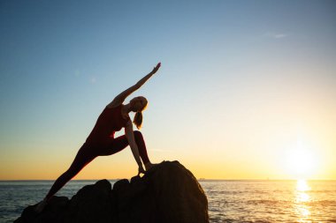 Gün batımında deniz sahilinde yoga yapan ve egzersiz yapan genç kadının silueti. 