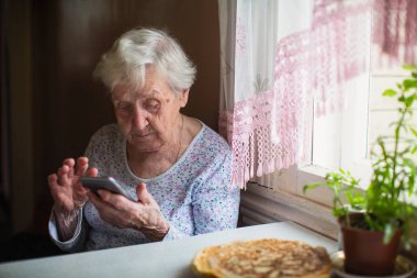 Yaşlı bir kadın evde elinde bir akıllı telefon ile oturur.