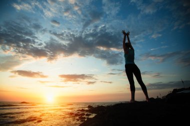 Genç kadın şafak sırasında deniz kıyısında yoga egzersizleri yapıyor.