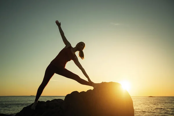 Woman silhouette doing yoga exercise on sea beach during surreal sunset.