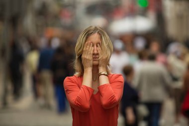 Young woman covering her face with her hands at standing in the middle of a crowded street. Panic attack in public place.  clipart