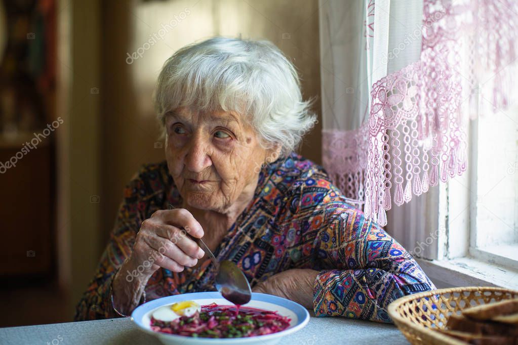 Elderly woman eating borsch soup sitting at the table in his home.
