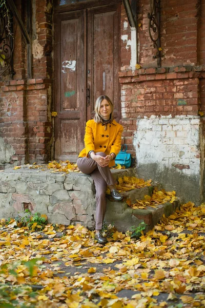 秋天的空气中 穿着黄色夹克 头戴蓝色布袋的女人坐在一座古旧房子的门廊上 地上长满了枫叶 — 图库照片