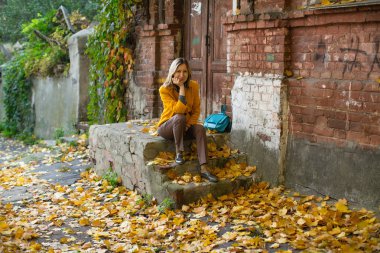 Attractive blonde woman in a yellow jacket poses in an autumn street with red and yellow foliage.