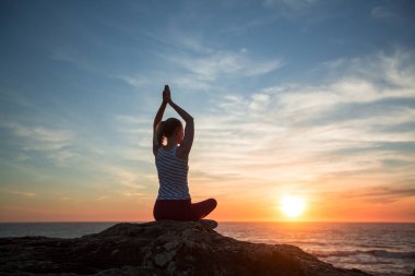 Genç yogacı kadının silueti okyanus sahilinde gün batımında meditasyon yapıyor..  