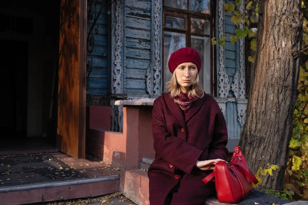 A sad woman in a coat sits on the porch of an vintage country house.