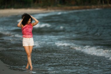 Melez bir kadın tropikal deniz sahilinde dalgaların kenarında yürüyor..