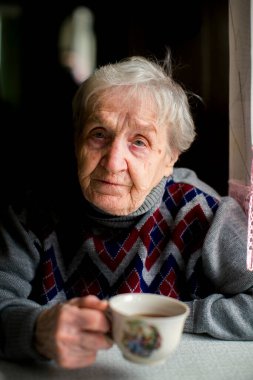 Yaşlı bir kadın elinde fincanla masada oturuyor..