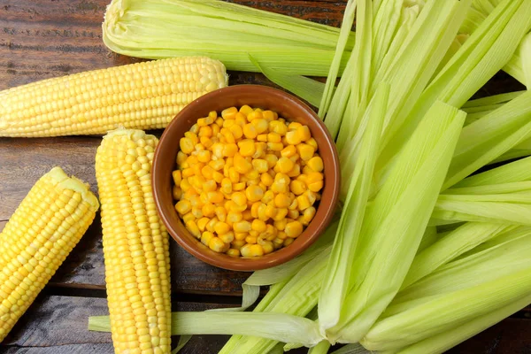 Raw corn kernels, inside ceramic bowl, next to corn on the cob on rustic wooden table. top view