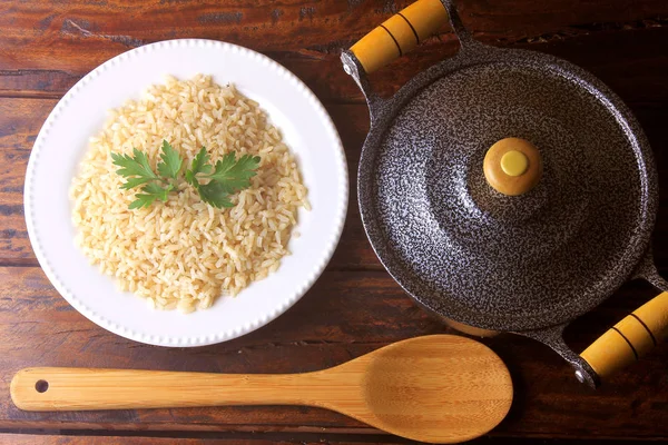 Arroz integral cozido no prato ao lado de panela e colher de bambu na cozinha em mesa de madeira rústica — Fotografia de Stock