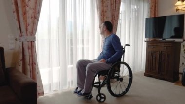 Üzgün genç adam evde pencereden bakarak tekerlekli sandalye veya bakım evi. Yavaş hareket 4k el hareketi
