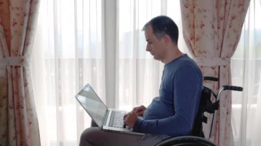 Genç adam dizüstü bilgisayarda evde pencerenin yanında çalışan tekerlekli sandalyede. Yavaş hareket 4k el hareketi