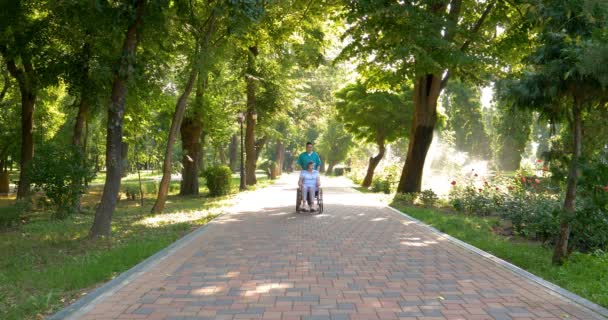 年轻的护士与坐在轮椅上的老妇人在公园里散步 度过了阳光灿烂的一天 — 图库视频影像