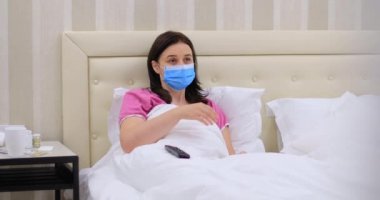 Tıbbi maskeli bir kadın karantinada evinde televizyon seyrediyor. Koronavirüs salgını sırasında izole edilmiş bir kadın. Dolly 4K çekti.
