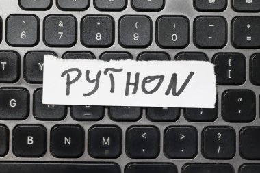 Kara klavyeye, bilgisayar bilimini ve programlamayı sembolize eden bir kağıt parçasına el yazısıyla yazılmış kelime PYTHON. Teknolojiyle ilgili projeler için uygun.