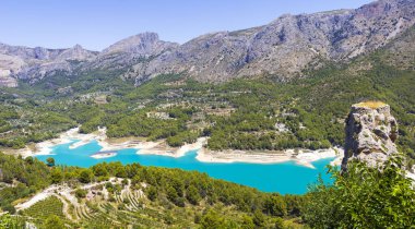 Bereketli yeşil ormanlarla çevrili turkuaz göl ve berrak mavi gökyüzünün altında yükselen dağlarla dolu Guadalest İspanya 'nın çarpıcı panoramik manzarası. Açık hava meraklıları için sakin bir manzara..