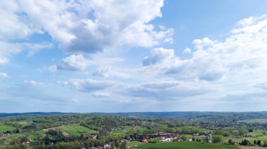 Kabarık bulutlarla dolu parlak mavi bir gökyüzü altında yuvarlanan tepelerin, yemyeşil ve dağınık köylerin yer aldığı pitoresk kırsal bir manzaranın havadan görünüşü. Doğa temalı projeler için ideal.