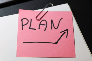 Üzerinde PLAN yazan canlı pembe yapışkan bir not ve üretkenlik temaları için ideal beyaz bir defter üzerine yerleştirilmiş büyüme ve organizasyonu sembolize eden yukarı doğru bir ok..