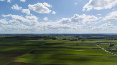 Tüylü beyaz bulutlarla benekli parlak mavi gökyüzü altında kahverengi toprak parçalarıyla yemyeşil alanlar. Bu pitoresk kırsal manzara tarım güzelliğini gözler önüne seriyor.