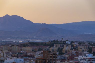 Alicante 'nin şehir manzarası ve gün batımında dağları gösteren manzarası. Ufuk çizgisi doğal güzellik ve tepelere karşı konut binaları sergiliyor..
