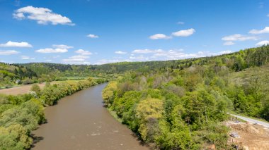 Parlak mavi gökyüzünün altında yemyeşil ağaçlarla çevrili dolambaçlı bir nehrin havadan görünüşü. Sakin manzara, ilkbaharda doğanın güzelliğini gözler önüne seriyor..