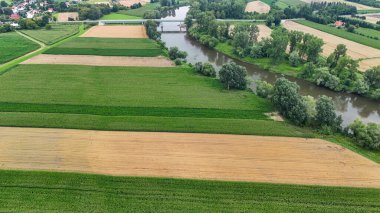 Yeşil yemyeşil tarlalar ve kırsal alandaki ağaçlar ve tarımsal arazilerle çevrili bir nehir manzarası..