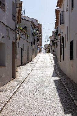 Altea İspanya - 17 Temmuz 2025: Altea 'da dar bir güneş ışığı sokağı geleneksel beyaz boyalı binalar ve kaldırım taşlı yollar sergiliyor ve canlı bir Akdeniz köyünde büyüleyici bir atmosfer yaratıyor..