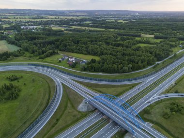 Yeşillik ve ormanlarla çevrili bir otoyol kavşağının havadan görünüşü. Bu sahne, kırsal bölgeleri gösteren mavi bir köprüyle yolların kesiştiği yeri gösteriyor..