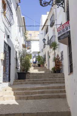 Altea İspanya - 17 Temmuz 2025 Altea 'da merdivenlerden çıkan bir adamla dar bir sokak. Beyaz binalar ve saksılarla çevrili büyüleyici bir atmosfer yaratıyorlar..