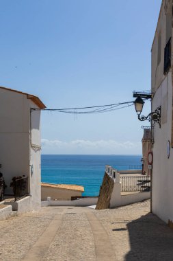 Altea Spain - 17 Temmuz 2025: Akdeniz 'e uzanan dar bir sokakta beyaz yıkanmış binalar ve açık mavi gökyüzü kıyı yaşamının özünü yansıtıyor.