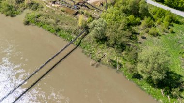 Sudan geçen ahşap bir köprünün bulunduğu yemyeşil yemyeşil bir nehir kıyısının havadan görünüşü. Bu sakin sahne doğanın güzelliğini ve sükunetini yansıtıyor..