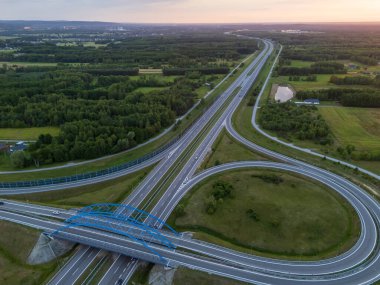 Günbatımında yeşil manzaralar ve yol ağlarının yer aldığı modern bir otoyol kavşağının havadan görünüşü. Ulaşım veya kentsel planlama temaları için ideal.