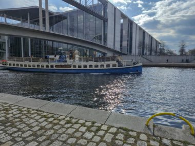 Berlin Almanya - 9 Nisan 2025: Modern mimarinin yakınlarındaki sakin sularda süzülen büyüleyici bir tekne, güneş ışığını yüzeyde yansıtıyor ve sakin bir manzara yaratıyor.