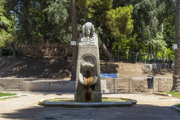 Alicante İspanya - 13 Temmuz 2025: Palmiye ağaçları ve yeşilliklerle çevrili sakin bir parkta bir tanrıça heykeli bulunan çarpıcı bir taş çeşme.