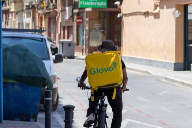 Alicante İspanya - 17 Temmuz 2025: Bir gıda dağıtım bisikletçisi Alicante 'de sakin bir caddede bisiklet sürüyor ve şehir yaşamındaki dağıtım hizmetlerinin yükselişini gösteriyor.