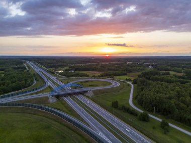 Gün batımında yemyeşil çayırlarla çevrili bir otoyol kavşağının havadan görüntüsü. Yollar, doğanın ve altyapının güzelliğini gözler önüne seren ufka çıkar..