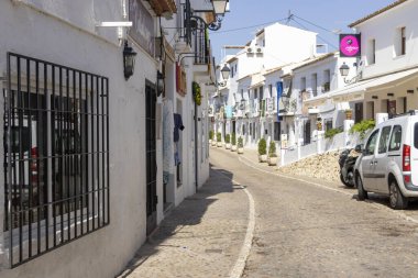 Altea İspanya - 17 Temmuz 2025: Altea 'da güneşli güneş ışığıyla yıkanmış büyüleyici beyaz boyalı binalar ve yerel dükkanlarla dolu tuhaf bir kaldırım taşı caddesi.